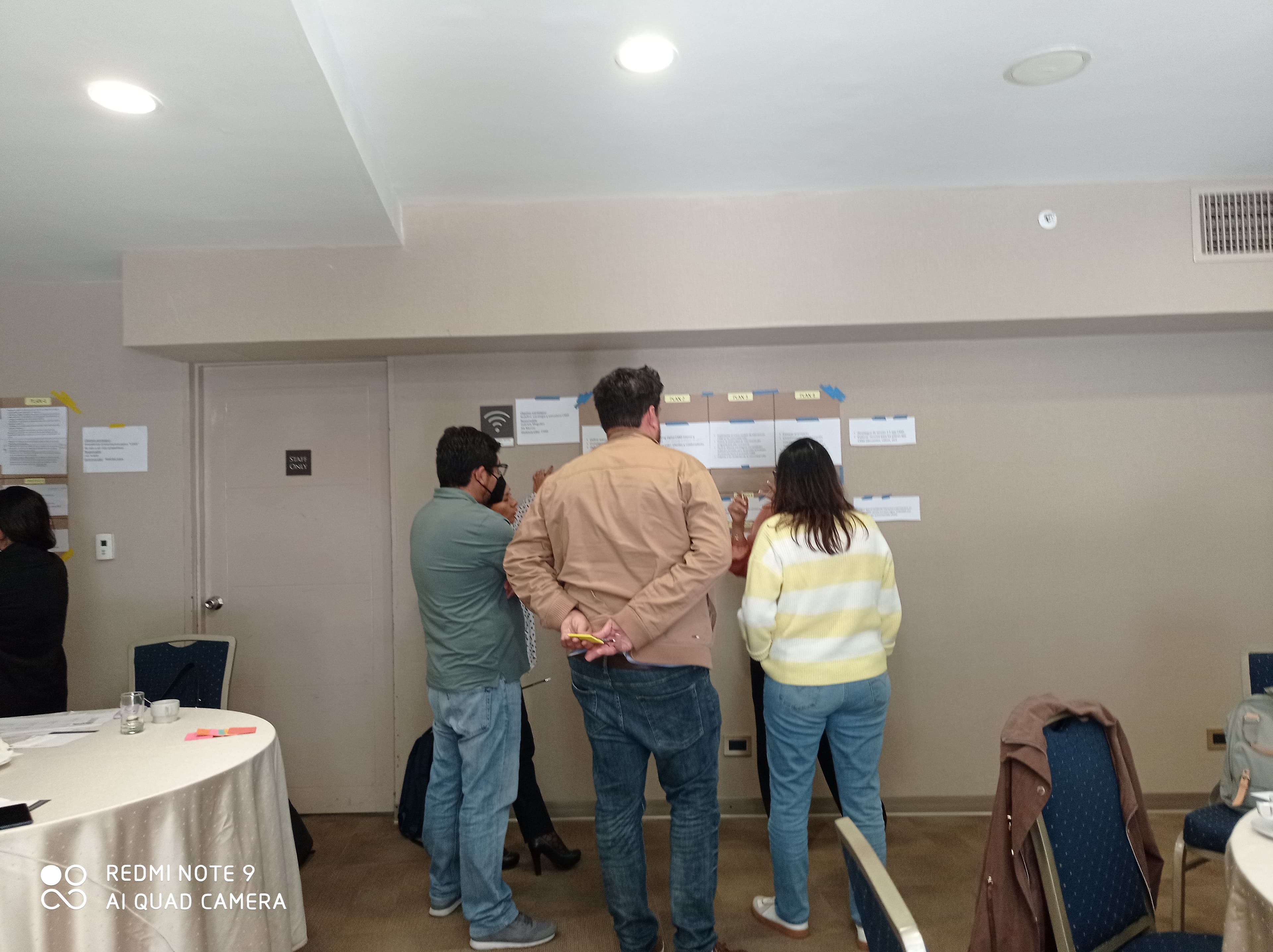 Grupo de personas analizando documentos pegados en la pared durante una sesión de trabajo colaborativo.