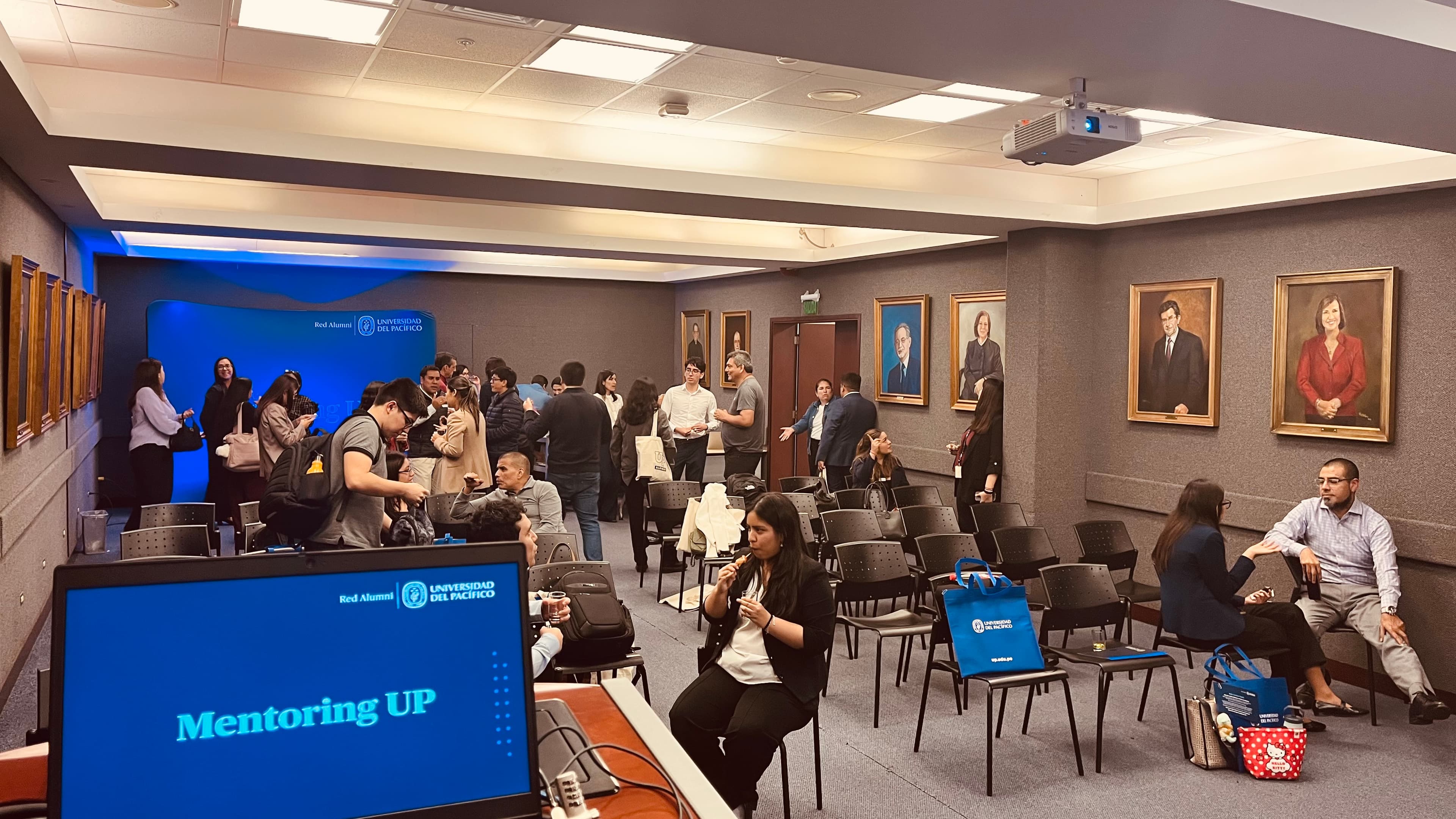 Asistentes conversando en un evento de Mentoring UP en una sala con retratos institucionales.